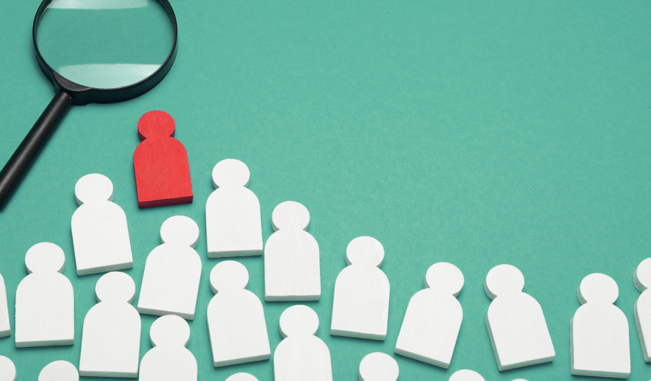 Magnifying glass highlighting a red wooden figure among white figures, representing the process of hiring the right Medical Virtual Assistant for healthcare practices.